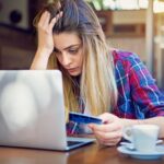 An investor touches their head in frustration while sitting at a cafe and holding a credit card while looking at a laptop.