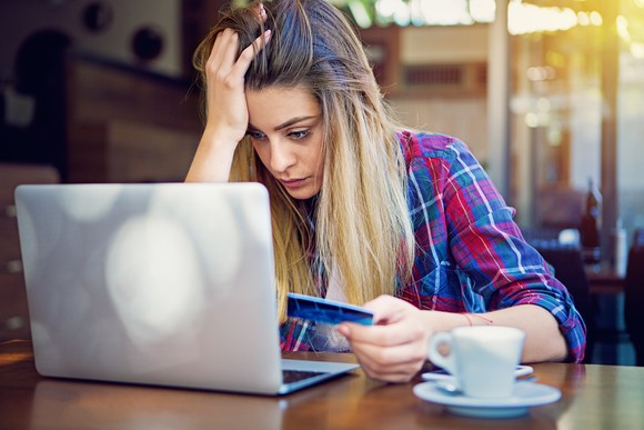 An investor touches their head in frustration while sitting at a cafe and holding a credit card while looking at a laptop.